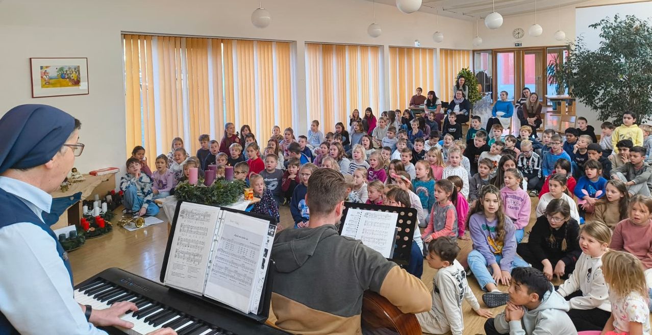 Eine Gruppe von Kindern und Erwachsenen in einem Raum mit gelben Jalousien, einige sitzen und einige stehen, lauschen einem Mann, der Klavier und Gitarre spielt, mit Notenblättern vor ihm.