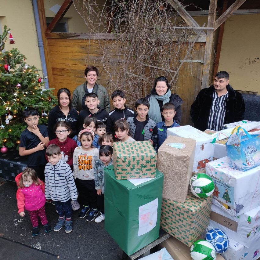 Eine Gruppe von Menschen, einschließlich Erwachsener und Kindern, steht vor einem Weihnachtsbaum und einem Stapel verpackter Geschenke. Sie scheinen für ein Foto zu lächeln.
