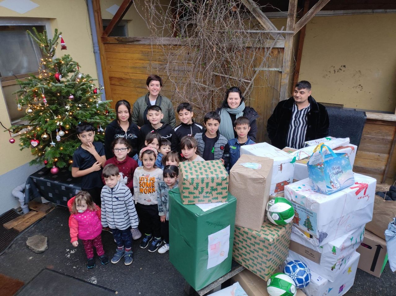 Eine Gruppe von Menschen, einschließlich Erwachsener und Kindern, steht vor einem Weihnachtsbaum und einem Stapel verpackter Geschenke. Sie scheinen für ein Foto zu lächeln.