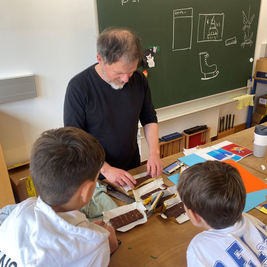 Ein älterer Mann unterrichtet zwei Kinder an einem Tisch. Sie untersuchen ein Stück Schokolade. Ein Tafel mit Zeichnungen ist im Hintergrund.