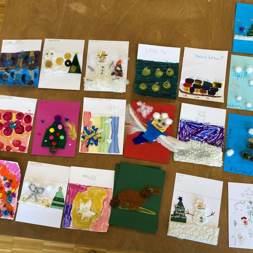 Ein hölzerner Tisch zeigt eine Sammlung festlicher Weihnachtskarten, die jeweils mit Elementen wie Schneemännern, Weihnachtsbäumen und bunten Ornamenten verziert sind. Die Karten variieren in Größe und Stil und zeigen verschiedene künstlerische Techniken und Materialien.