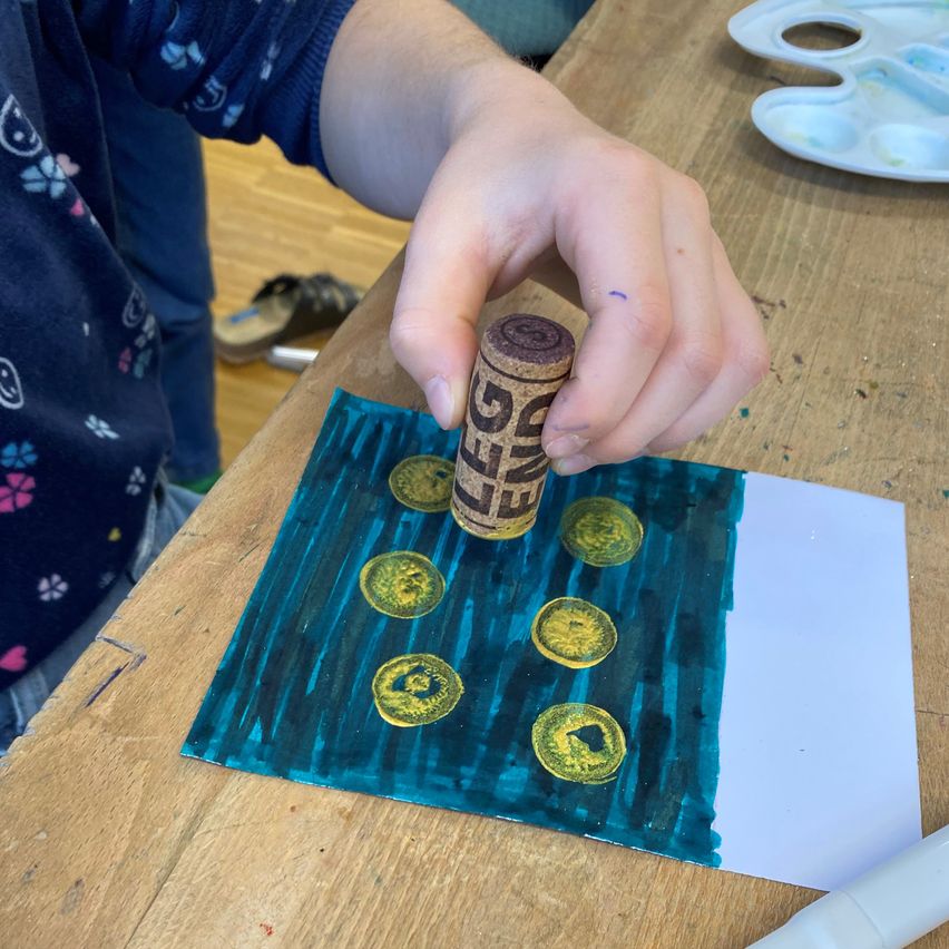 Ein Kind hält einen Korken mit einem Muster und wendet ihn auf ein Papier mit bemaltem Hintergrund und mehreren gelben Münzen an.