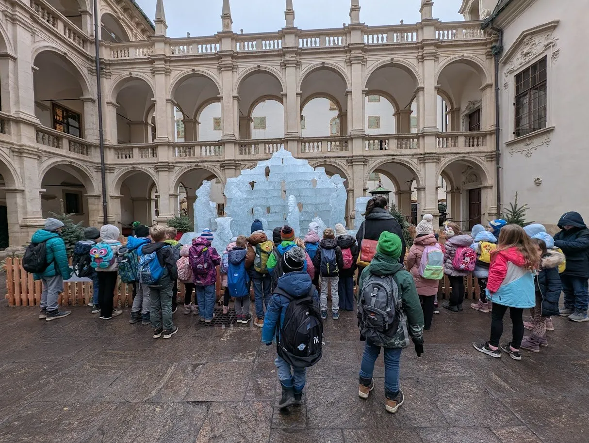 Eine Gruppe von Kindern in Winterkleidung und Rucksäcken steht um eine Eisskulptur herum auf einem Freihof eines historischen Gebäudes.
