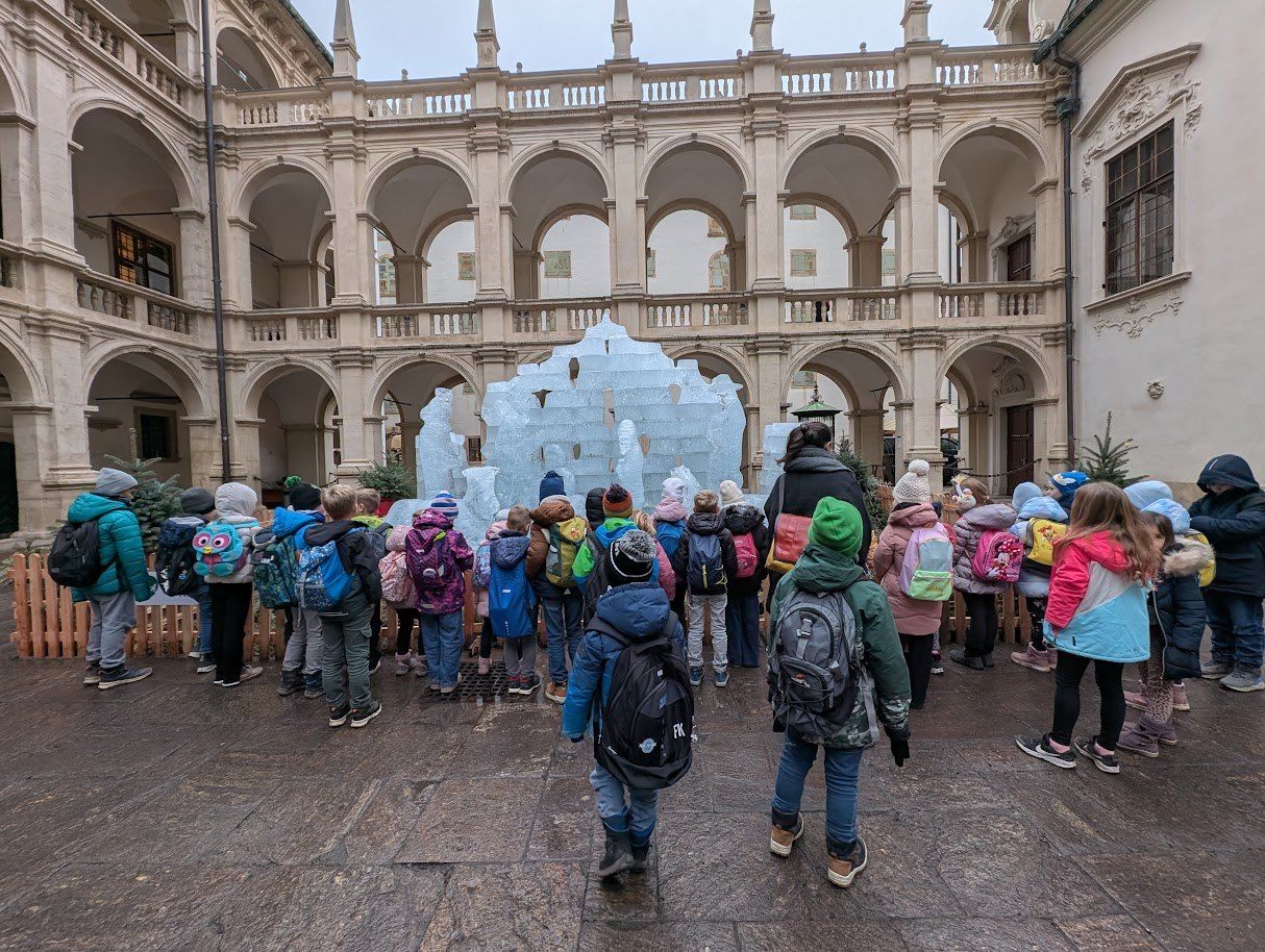 Eine Gruppe von Kindern in Winterkleidung und Rucksäcken steht um eine Eisskulptur herum auf einem Freihof eines historischen Gebäudes.
