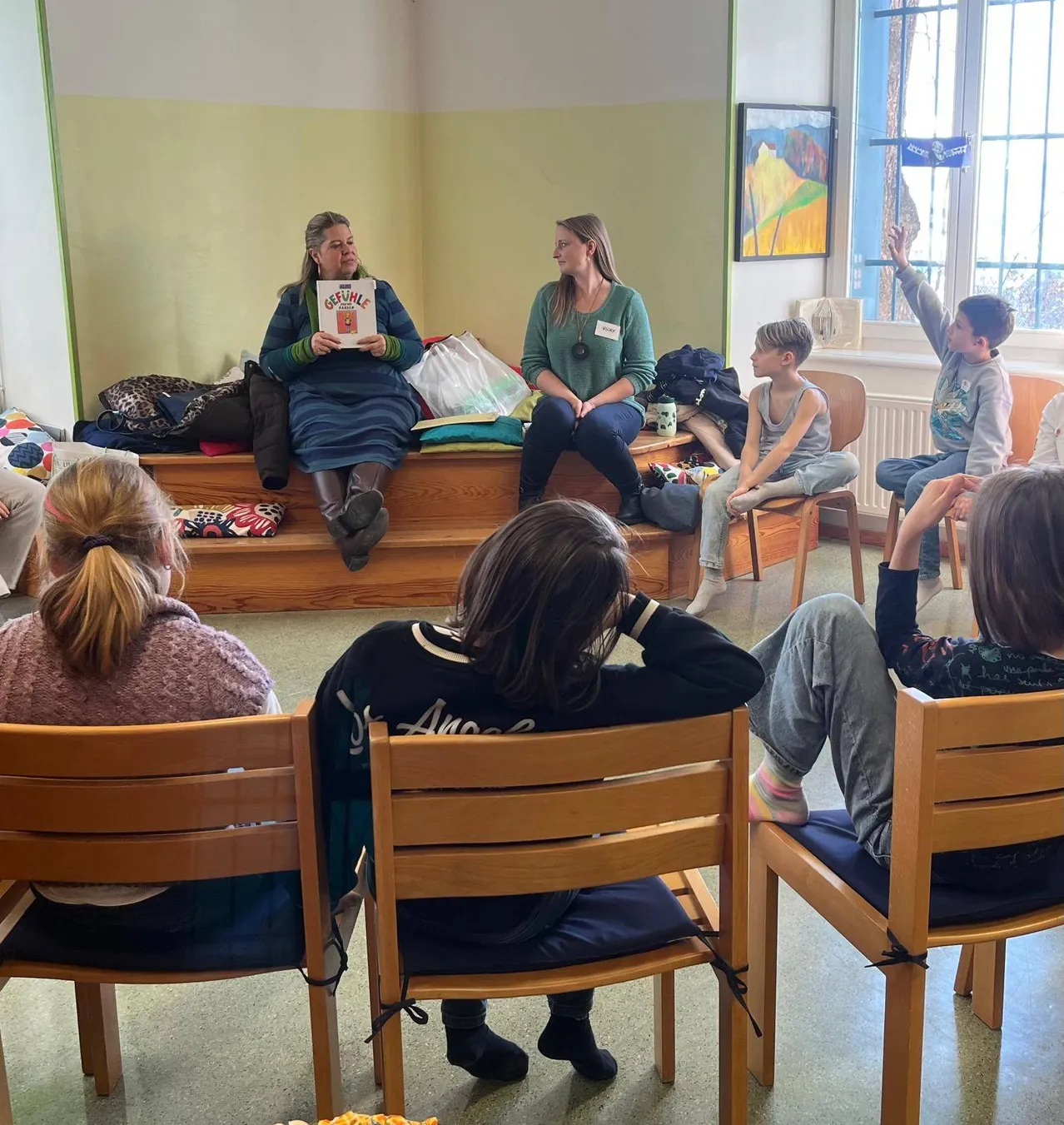 Eine Gruppe von Menschen sitzt in Stühlen in einem Raum. Zwei Frauen sitzen auf einer Bank und halten Bücher. Zwei Kinder sitzen auf Stühlen mit erhobenen Händen. Ein Gemälde ist an der Wand.