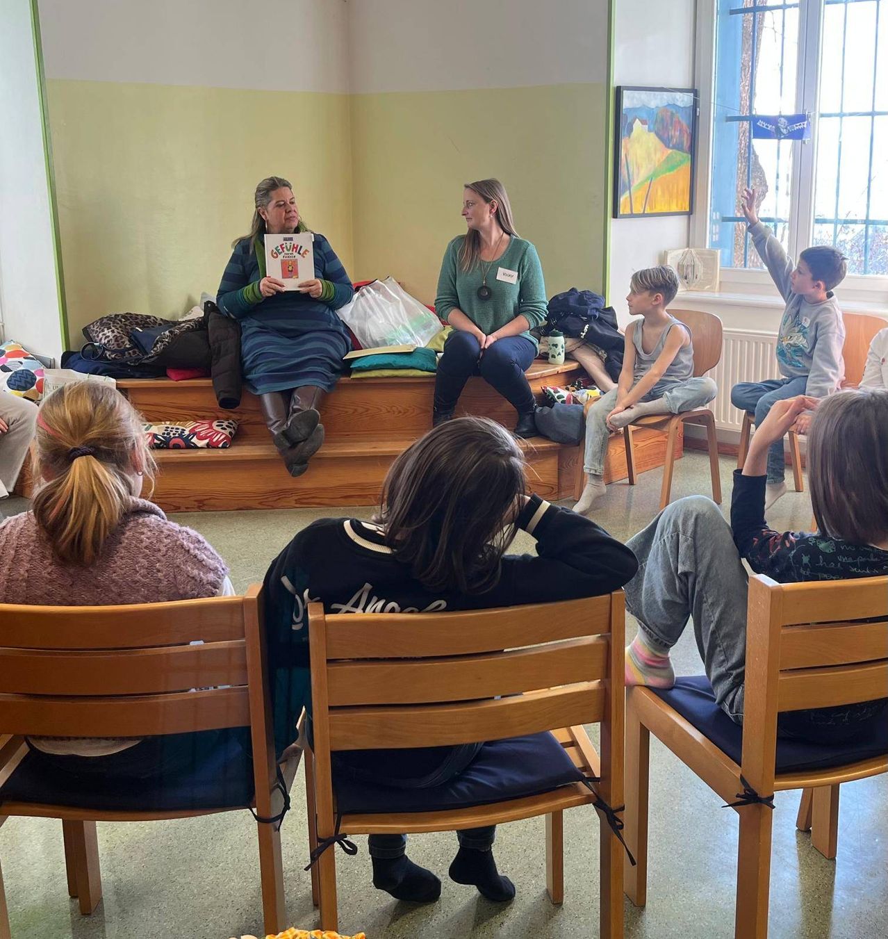 Eine Gruppe von Menschen sitzt in Stühlen in einem Raum. Zwei Frauen sitzen auf einer Bank und halten Bücher. Zwei Kinder sitzen auf Stühlen mit erhobenen Händen. Ein Gemälde ist an der Wand.