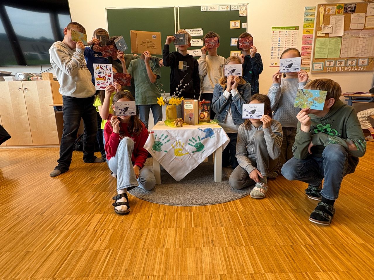 Eine Gruppe von Kindern posiert mit Büchern und dekorierten Papiermasken, einige sitzend und einige stehend um einen Tisch mit einer Vase mit Blumen herum. Sie sind in einem Klassenzimmer mit einem Bulletin Board im Hintergrund.