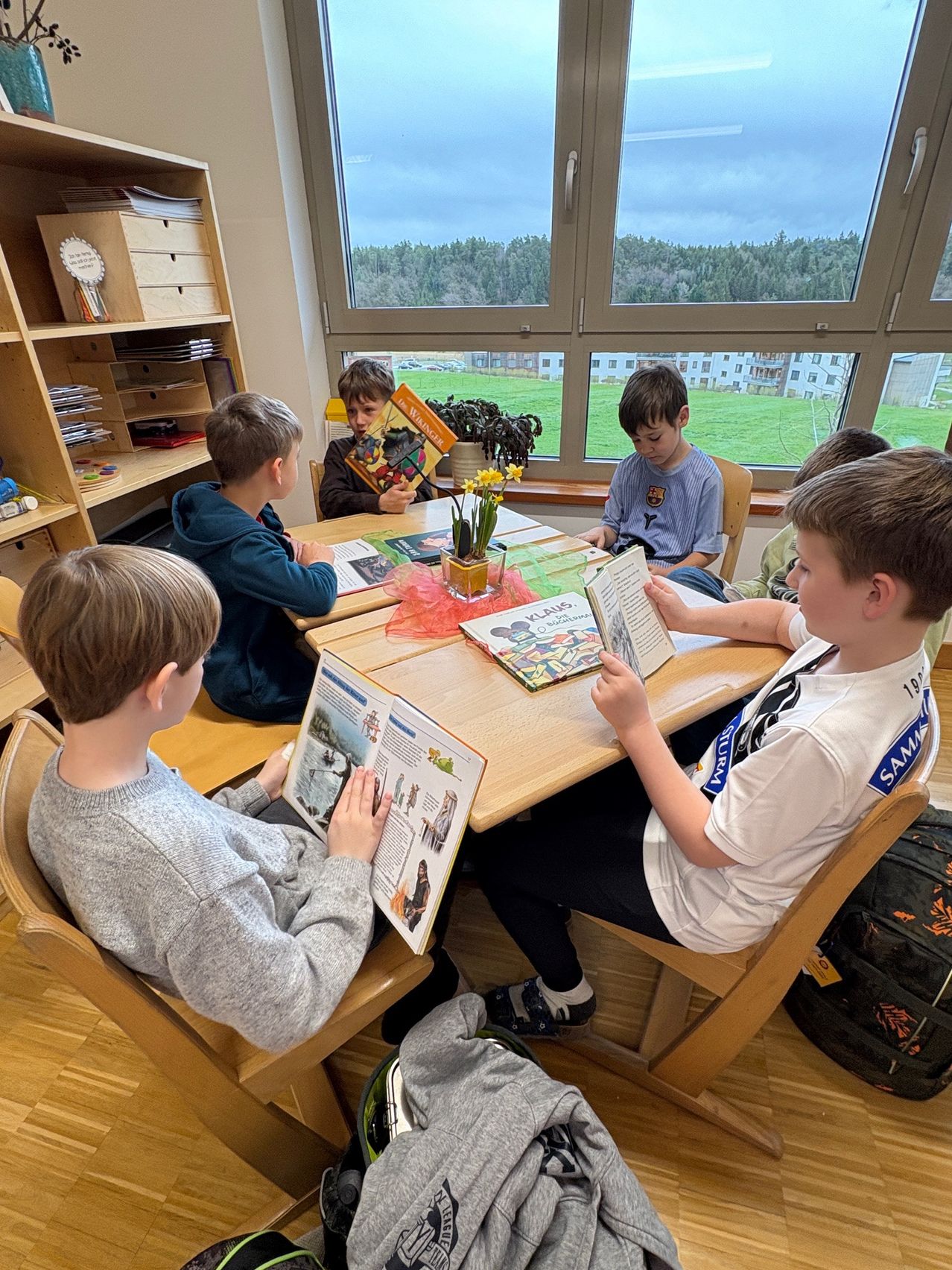 Eine Gruppe von Jungen sitzt um einen Tisch in einem Klassenzimmer herum und liest Bücher. Sie sind von Holzstühlen und Regalen mit Büchern umgeben. Das Zimmer hat große Fenster mit Blick auf ein grünes Feld und ein Gebäude.
