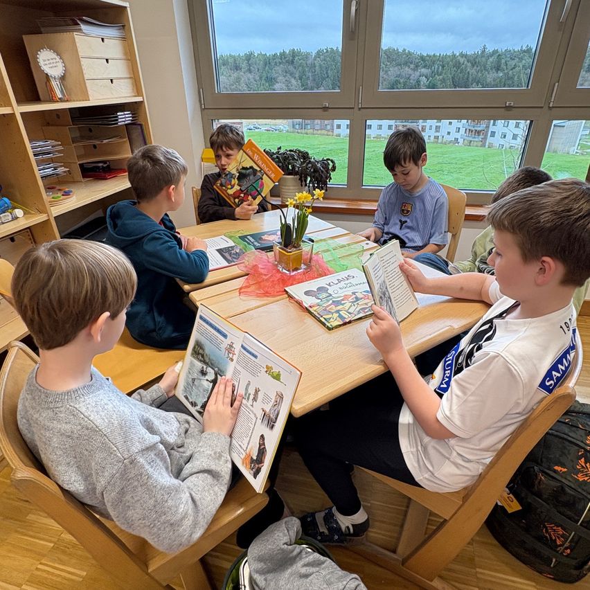 Eine Gruppe von Jungen sitzt um einen Tisch in einem Klassenzimmer herum und liest Bücher. Sie sind von Holzstühlen und Regalen mit Büchern umgeben. Das Zimmer hat große Fenster mit Blick auf ein grünes Feld und ein Gebäude.