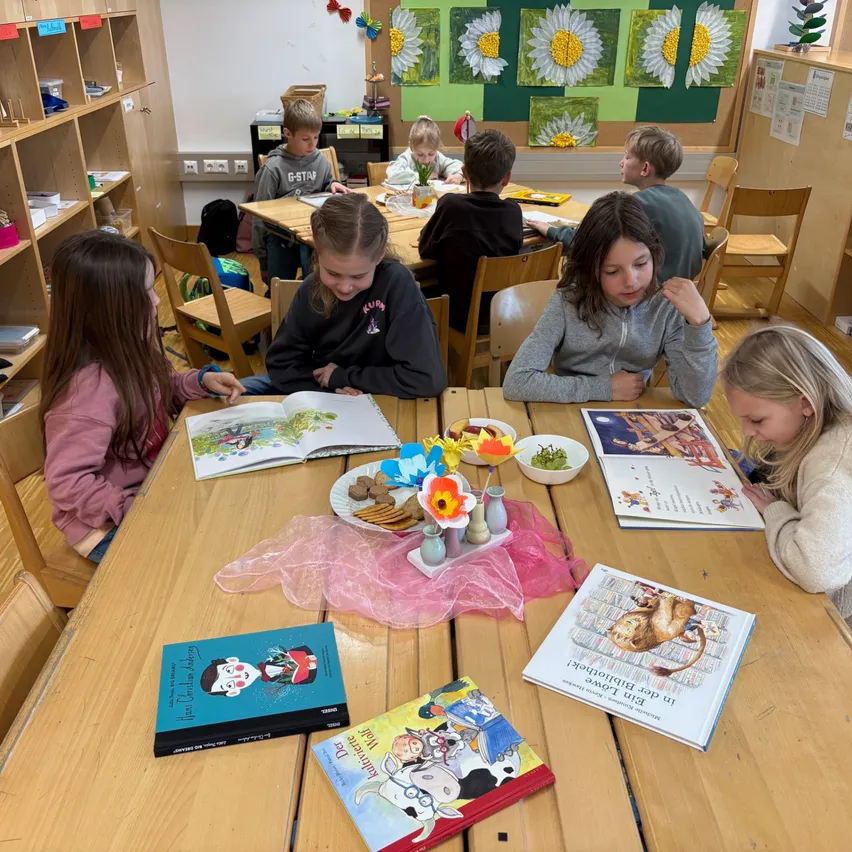 Ein Klassenzimmer mit Kindern, die um Tische herum sitzen. Bücher und Kekse sind auf den Tischen, und ein Mädchen schaut in ein Buch. Der Raum hat Regale mit verschiedenen Gegenständen.