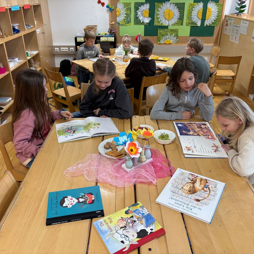 Ein Klassenzimmer mit Kindern, die um Tische herum sitzen. Bücher und Kekse sind auf den Tischen, und ein Mädchen schaut in ein Buch. Der Raum hat Regale mit verschiedenen Gegenständen.