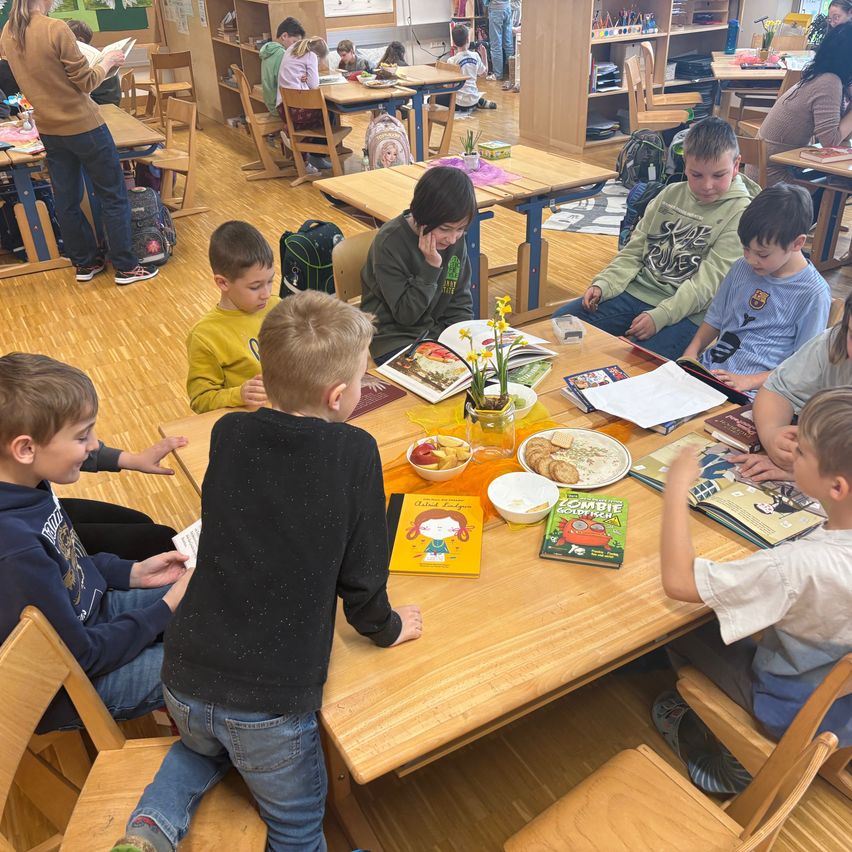 In einem Klassenzimmer sitzen Kinder um einen Tisch herum, einige lesen Bücher, während andere sich unterhalten. Ein Tisch im Vordergrund hat Bücher und Snacks, eine Frau steht daneben und liest.