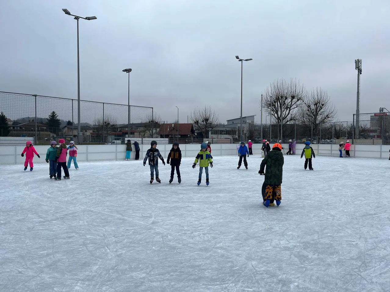 Kinder rutschen auf einer Eisbahn im Freien, andere beobachten und ein Gebäude im Hintergrund.