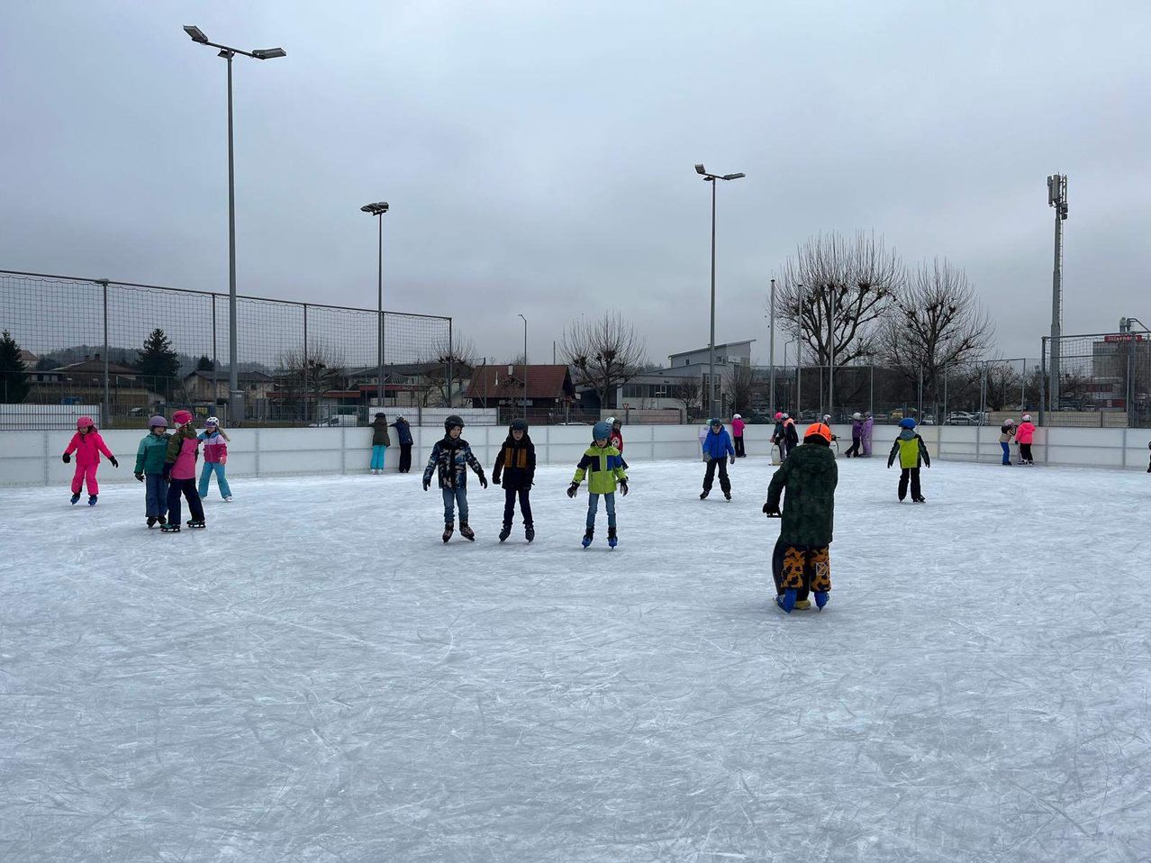 Kinder rutschen auf einer Eisbahn im Freien, andere beobachten und ein Gebäude im Hintergrund.