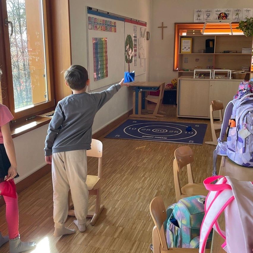 Ein junger Junge steht in einem Klassenzimmer und zeigt auf einen blauen Gegenstand über ihm. Ein weiteres Kind steht neben ihm. Der Raum hat Holzböden, ein Fenster und eine Tafel. Es gibt Stühle, einen Schreibtisch und ein Bücherregal.