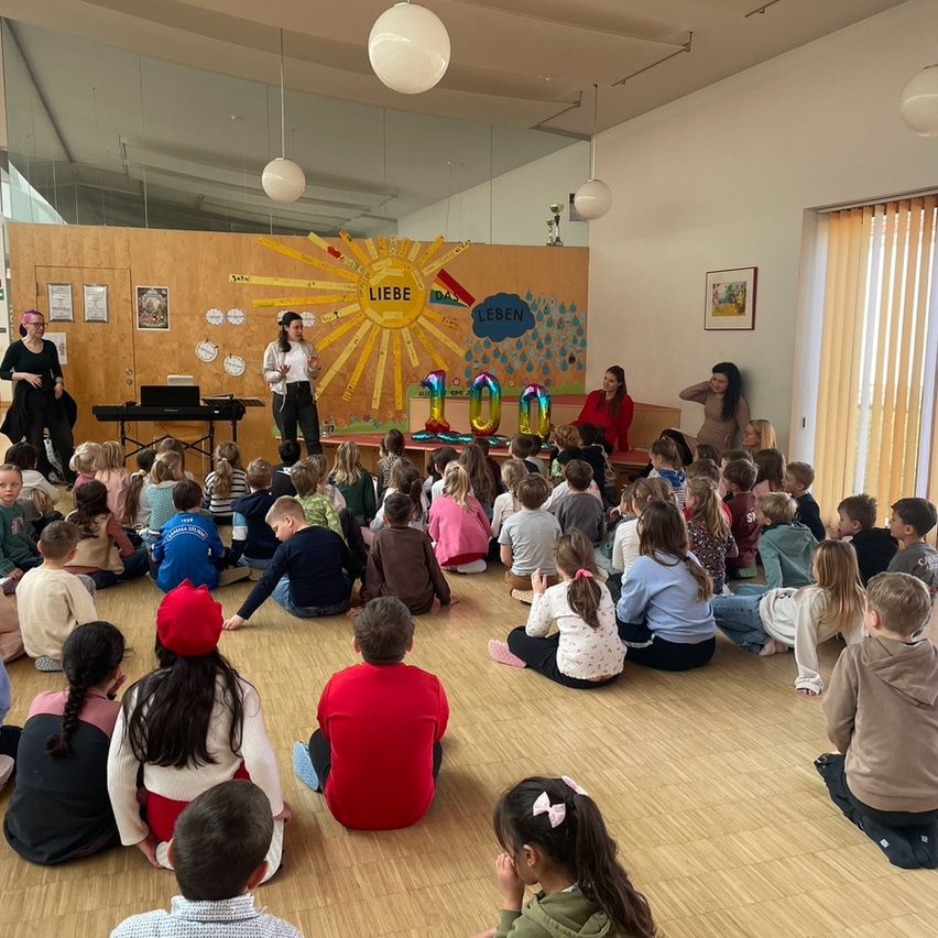 Kinder im Kreis lauschen einer Frau auf der Bühne mit einem Hintergrund aus Sonne und Ballonen. Eine andere Frau spielt Klavier.