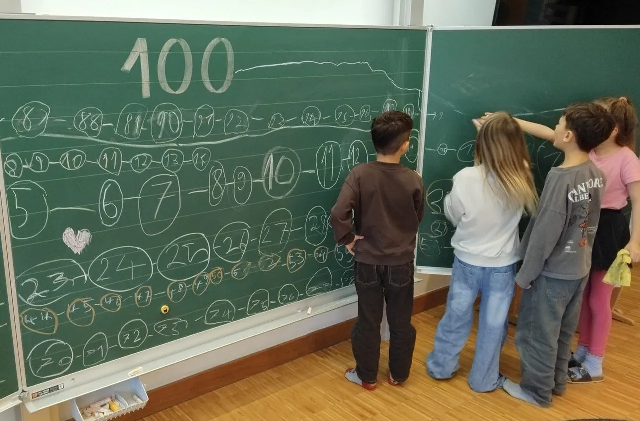 Zwei Kinder schauen auf eine Tafel mit Zahlen und mathematischen Gleichungen. Ein Junge trägt Braun, und ein Mädchen trägt Blau.