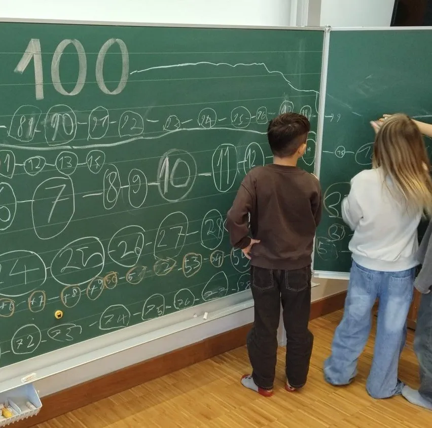 Zwei Kinder schauen auf eine Tafel mit Zahlen und mathematischen Gleichungen. Ein Junge trägt Braun, und ein Mädchen trägt Blau.