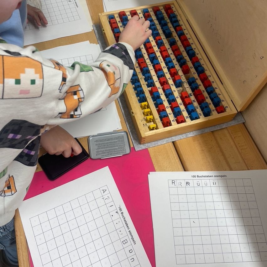 Ein Kind benutzt eine Holzbox mit verschiedenen farbigen Blöcken, um Buchstaben auf Papier zu stempeln. Die Box hat ein Raster mit Buchstaben, und das Kind hält einen Stempel.