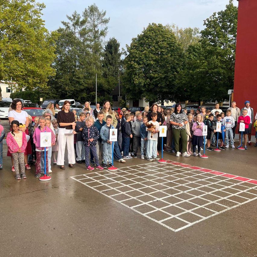Eine Gruppe von Kindern und Erwachsenen steht in einer Reihe auf einem nassen Bürgersteig und hält Schilder. Sie sind von Bäumen und geparkten Autos umgeben.