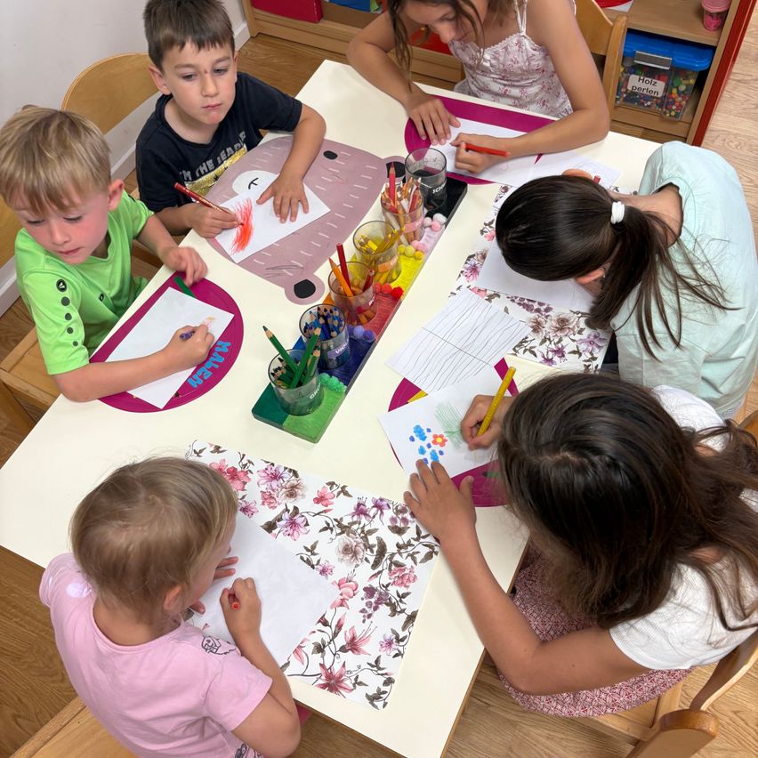 Eine Gruppe von Kindern in einem Klassenzimmer, die an einer Kunstaktivität teilnehmen. Sie sitzen um einen Tisch herum, jeder mit Papier und farbigen Stiften. Der Tisch ist mit verschiedenen Malutensilien gefüllt.