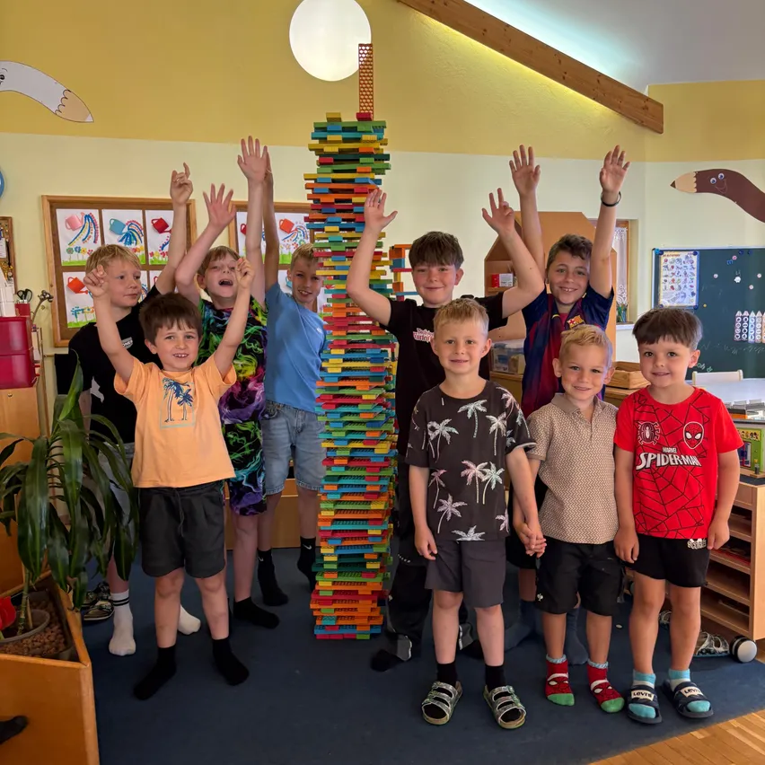 Eine Gruppe von Jungen steht in einem Klassenzimmer und posiert mit einem hohen Turm aus bunten Bausteinen. Sie haben ihre Hände in der Luft erhoben.