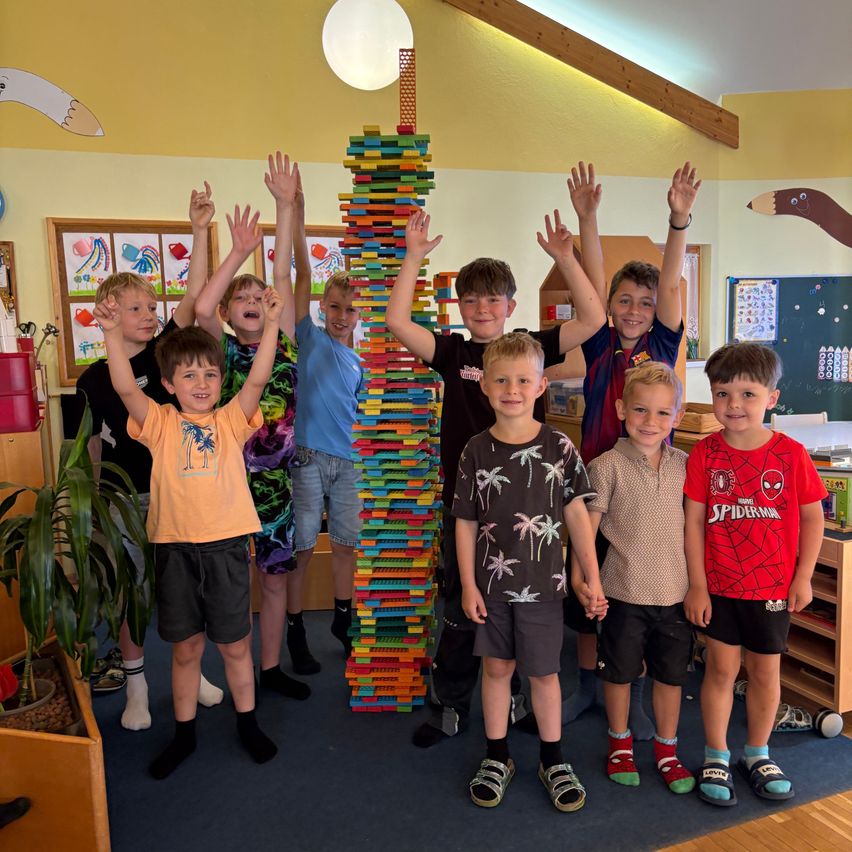 Eine Gruppe von Jungen steht in einem Klassenzimmer und posiert mit einem hohen Turm aus bunten Bausteinen. Sie haben ihre Hände in der Luft erhoben.