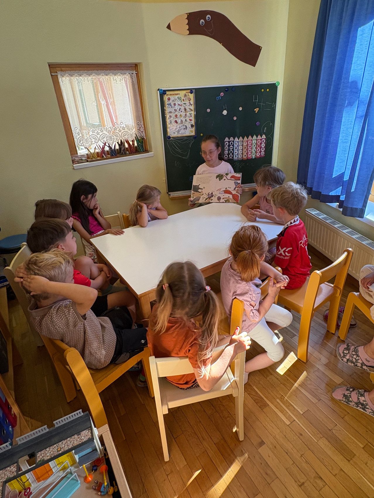 Eine Gruppe von Kindern sitzt um einen Tisch herum und hört einer Frau zu, die ein Buch liest. Einige Kinder schauen in das Buch, andere schauen woanders hin. Dahinter befindet sich eine Tafel mit Zeichnungen.