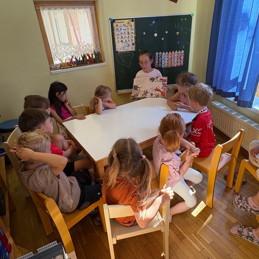 Eine Gruppe von Kindern sitzt um einen Tisch herum und hört einer Frau zu, die ein Buch liest. Einige Kinder schauen in das Buch, andere schauen woanders hin. Dahinter befindet sich eine Tafel mit Zeichnungen.