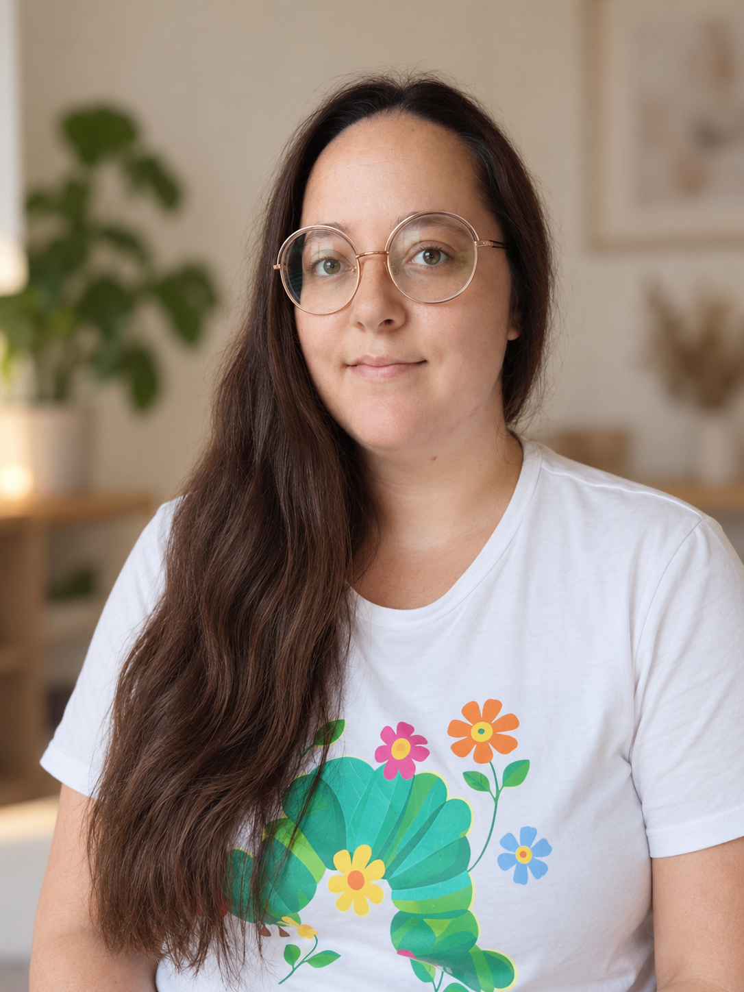 Eine Frau mit langen braunen Haaren, die eine Brille und ein weißes T-Shirt mit einem farbenfrohen Blumendesign trägt.