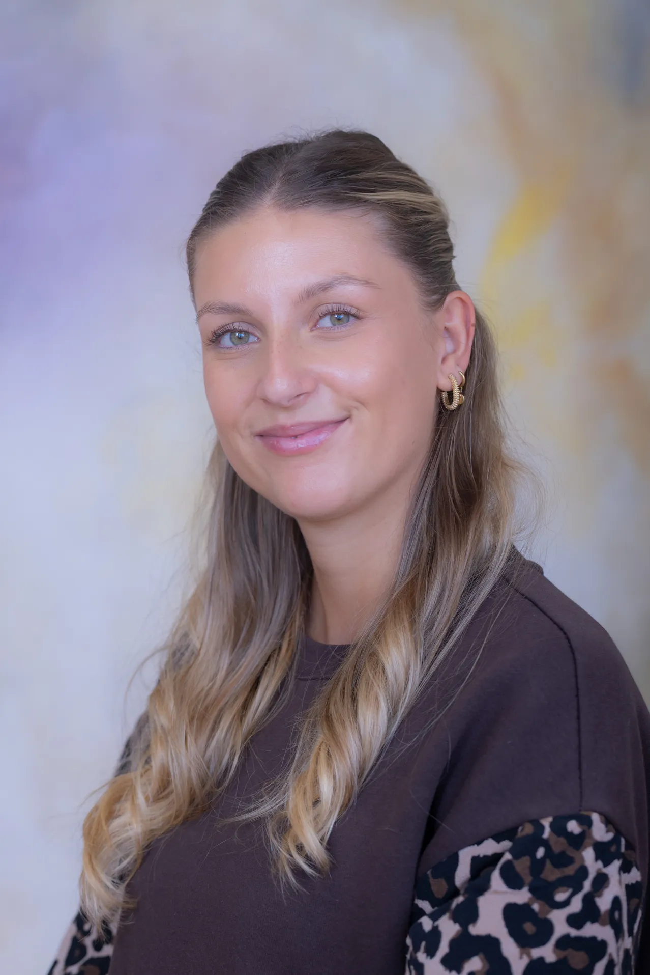 Eine Frau mit langen blonden Haaren, die ein dunkelbraunes T-Shirt und goldene Creolen trägt, lächelt in die Kamera.