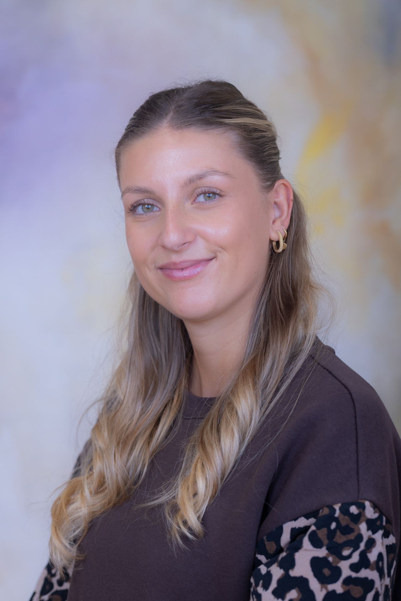 Eine Frau mit langen blonden Haaren, die ein dunkelbraunes T-Shirt und goldene Creolen trägt, lächelt in die Kamera.