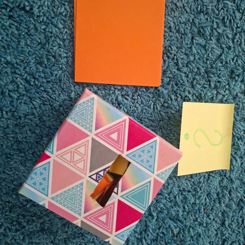 Eine bunte Box mit einem Dreiecksmuster ist geöffnet und zeigt ein kleines orangefarbenes Stück Papier im Inneren. Ein gelber Haftnotiz mit einem grünen Fragezeichen befindet sich neben der Box auf einem blauen Teppich.