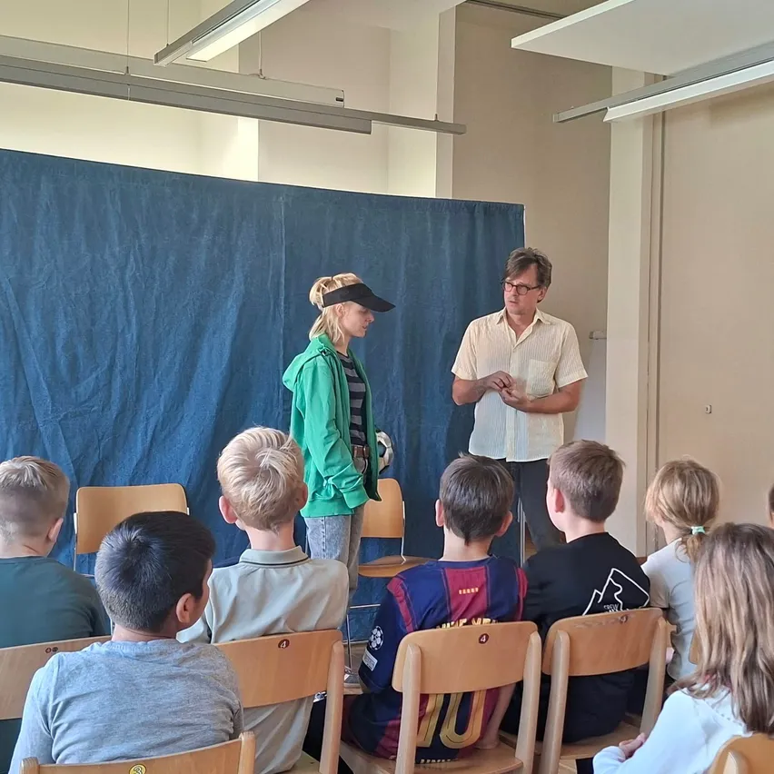 Eine Gruppe von Kindern sitzt in einem Klassenzimmer auf Stühlen. Zwei Erwachsene stehen vor ihnen, einer davon hält ein Stück Papier.