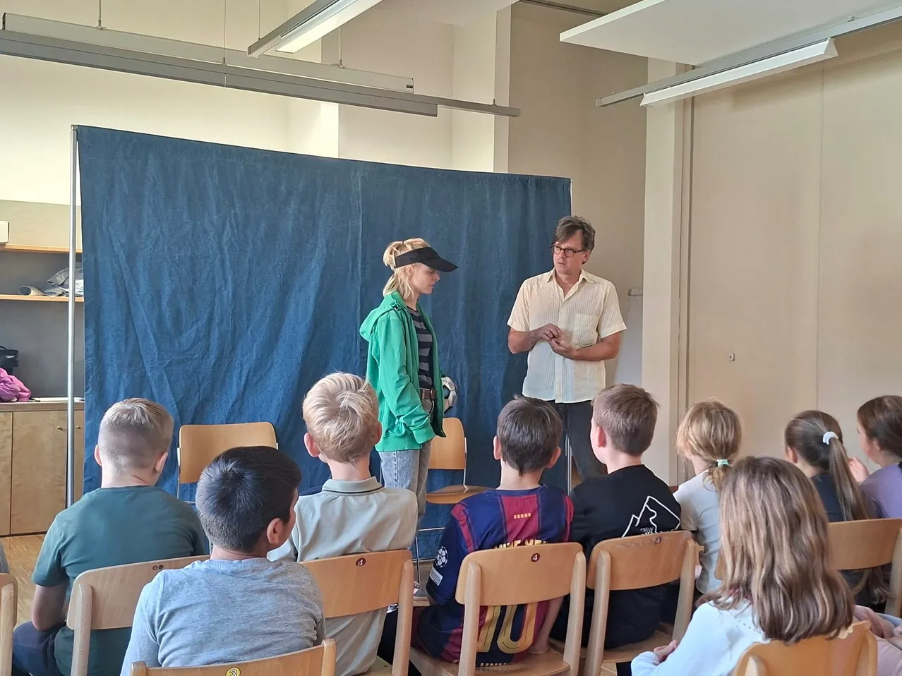 Eine Gruppe von Kindern sitzt in einem Klassenzimmer auf Stühlen. Zwei Erwachsene stehen vor ihnen, einer davon hält ein Stück Papier.