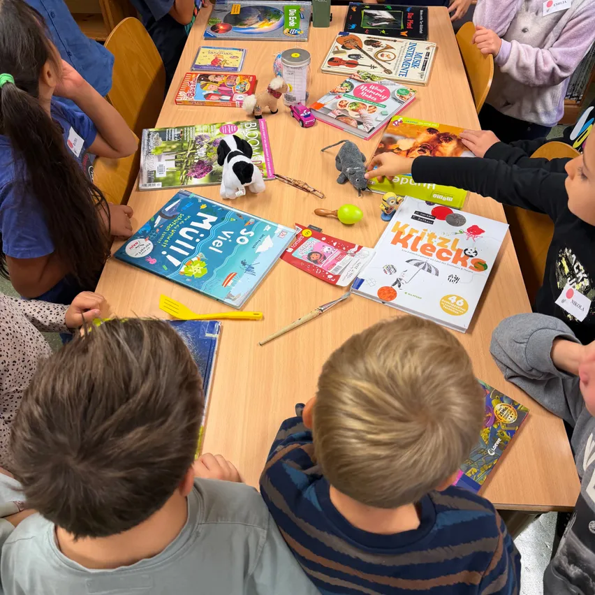 Eine Gruppe von Kindern sitzt in einer Bibliothek um einen Tisch herum und schaut sich Bücher und Spielzeug an.