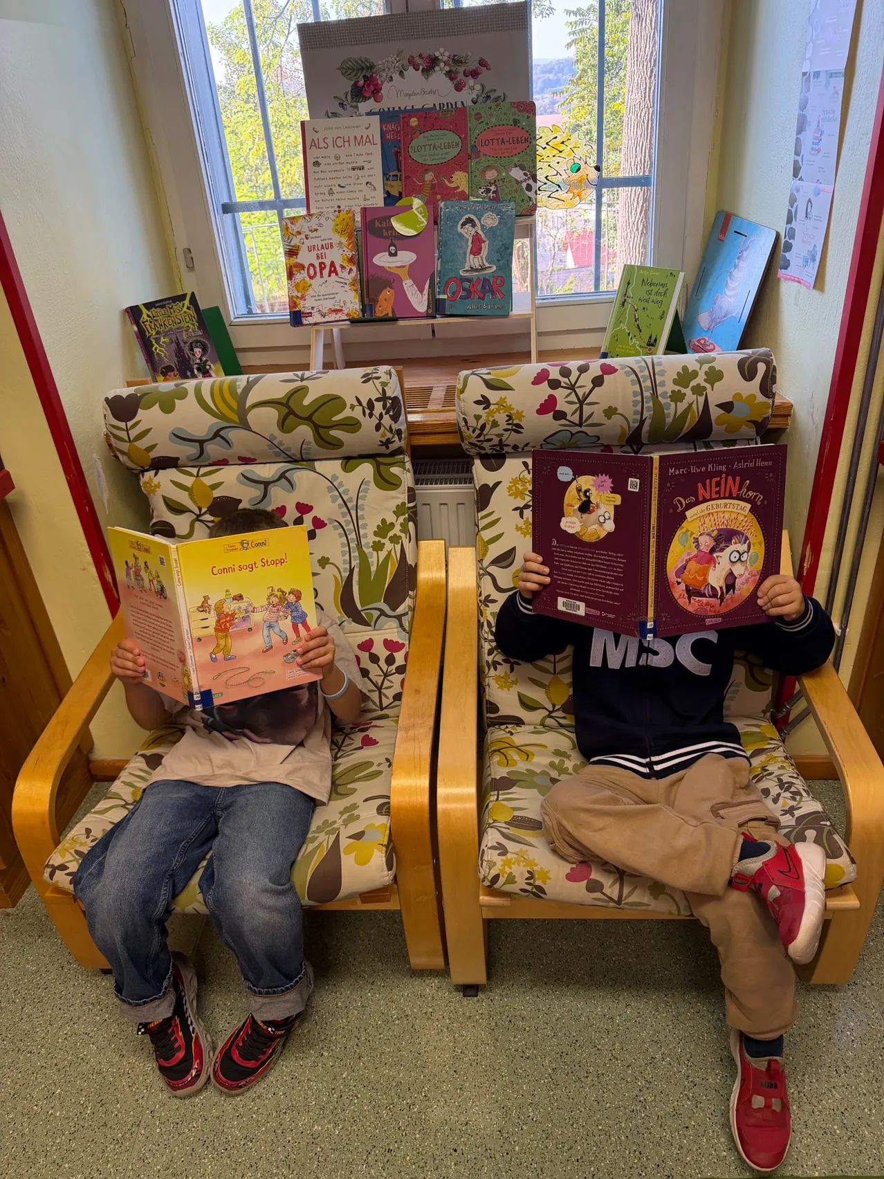 Zwei Kinder sitzen in Stühlen und lesen Bücher. Eines liest ein Buch mit dem Titel 'Conni auf Stapff' und das andere liest ein Buch mit dem Titel 'Die NEIN-Abenteuer'. Sie sitzen in einem Raum mit einem Fenster hinter ihnen.