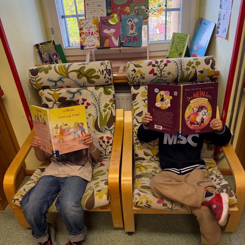 Zwei Kinder sitzen in Stühlen und lesen Bücher. Eines liest ein Buch mit dem Titel 'Conni auf Stapff' und das andere liest ein Buch mit dem Titel 'Die NEIN-Abenteuer'. Sie sitzen in einem Raum mit einem Fenster hinter ihnen.
