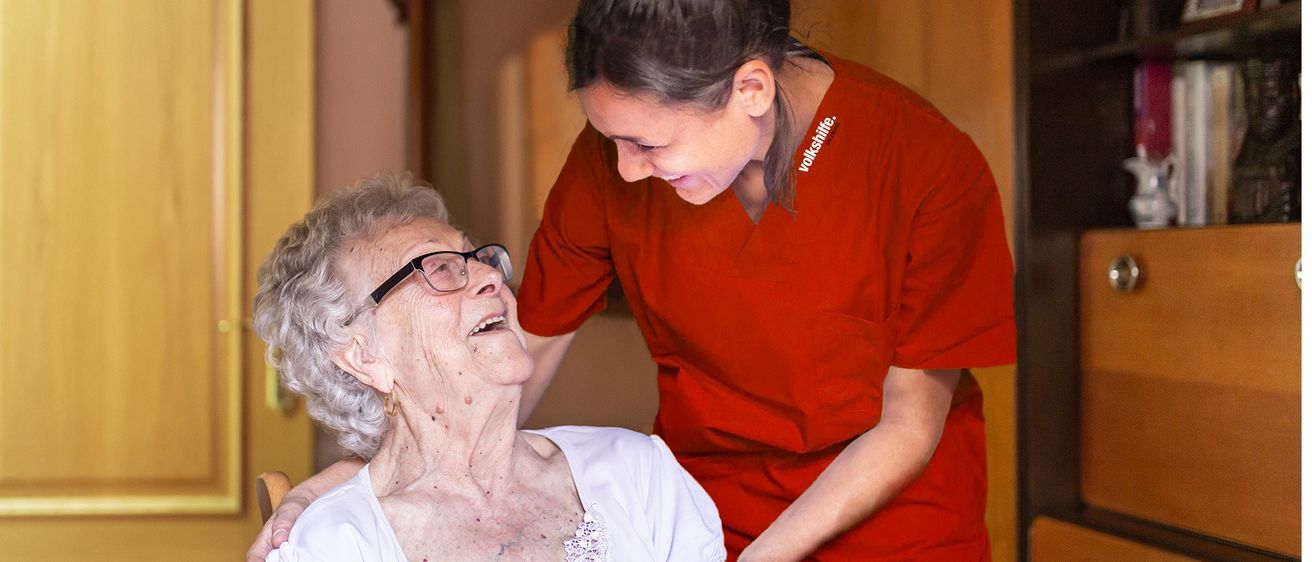 Eine ältere Frau lacht, während eine junge Frau mit Brille hinter ihr steht und ihre Schultern hält. Die junge Frau trägt ein rotes Shirt.