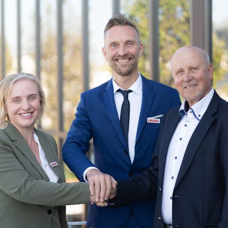 Drei Personen stehen nebeneinander und lächeln. Der Mann links trägt einen Anzug und eine Krawatte. Der Mann rechts trägt einen Blazer.