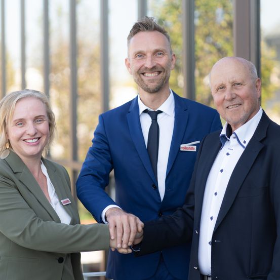 Drei Personen stehen nebeneinander und lächeln. Der Mann links trägt einen Anzug und eine Krawatte. Der Mann rechts trägt einen Blazer.