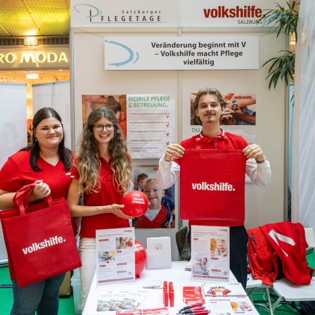 Drei Personen in roten Shirts stehen vor einem Stand. Die beiden Frauen halten rote Taschen und ein Mann hält eine rote Tasche mit dem Wort 'volkshilfe'. Das Banner über ihnen sagt 'Veränderung beginnt mit V'.