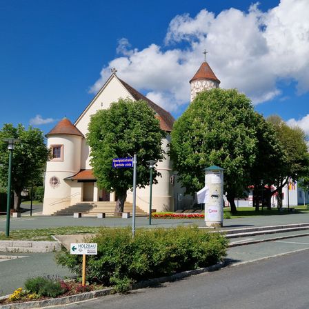 Eine weiße Kirche mit einem braunen Turm steht vor einem blauen Himmel. Ein Schild weist auf Holzbau hin. Bäume und Büsche stehen davor.