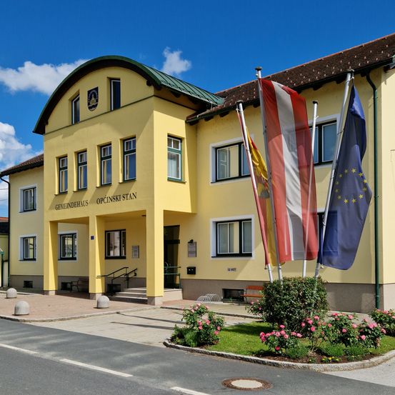 Das gelbe Gebäude mit grünem Dach ist ein Gemeindehaus, das Flaggen und einen Garten mit rosa Blumen vor sich hat.