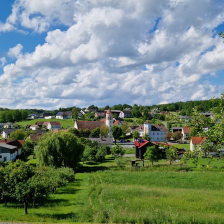 Ein malerisches Dorf in einem Tal mit einem Kirchturm, der über den Dächern aufragt, unter einem teilweise bewölkten Himmel.