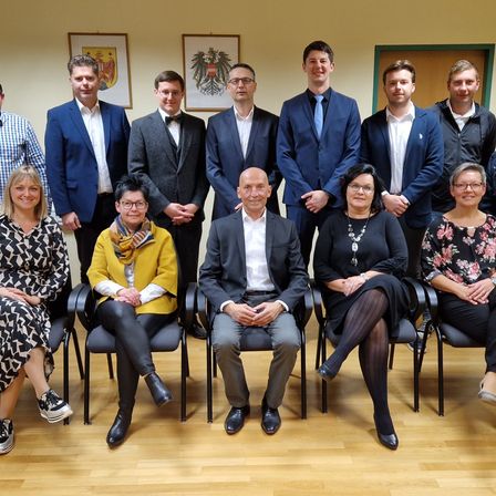 Eine Gruppe von Menschen posiert für ein Foto und trägt formelle Kleidung. Ein Mann in der Mitte sitzt auf einem Stuhl, und andere stehen hinter ihm. Der Raum hat Holzböden und gelbe Wände. Es hängen zwei Rahmen an der Wand.