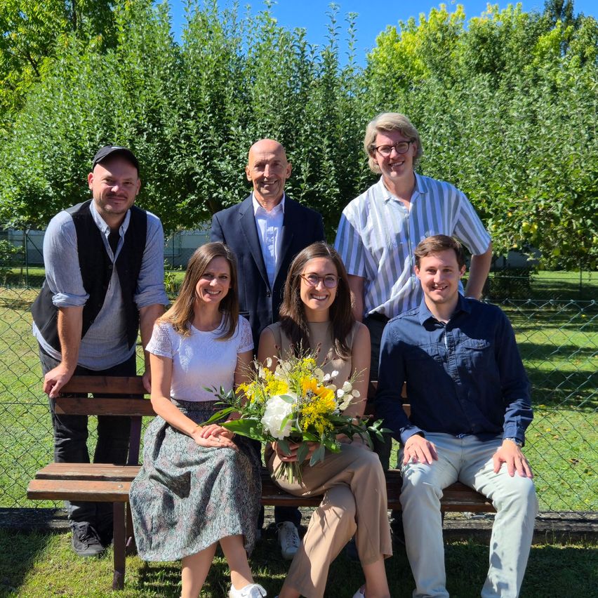 Eine Gruppe von sechs Personen, darunter zwei Frauen, posiert für ein Foto auf einer Bank in einem Garten mit Bäumen und einem Zaun im Hintergrund.