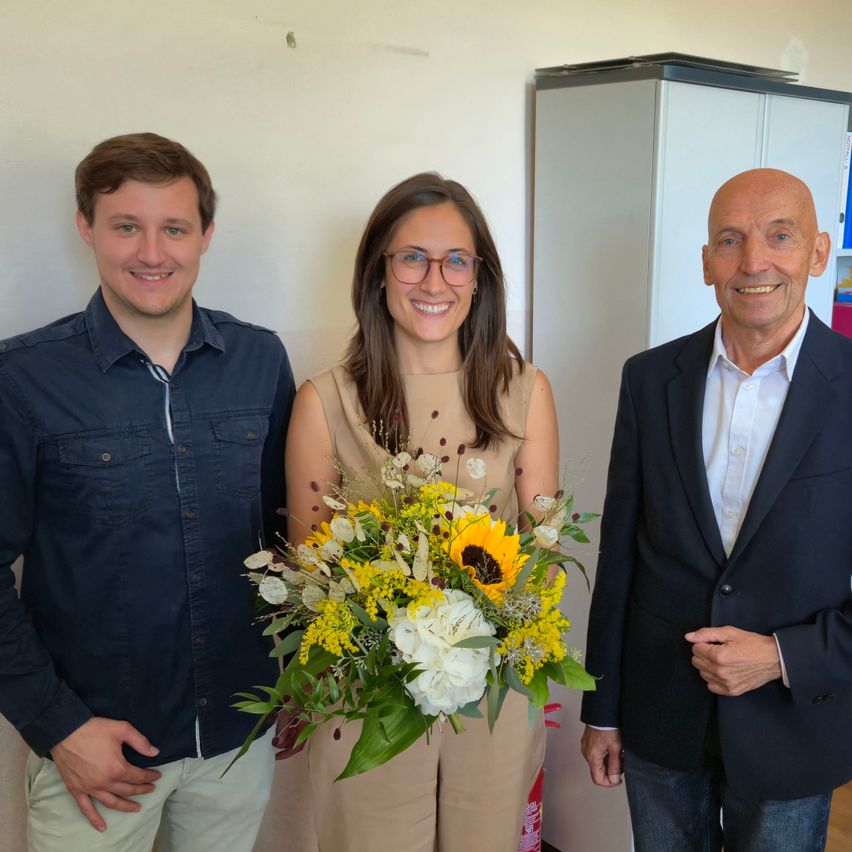 Drei Personen, zwei Männer und eine Frau, stehen lächelnd vor einer weißen Wand. Die Frau hält einen Blumenstrauß.