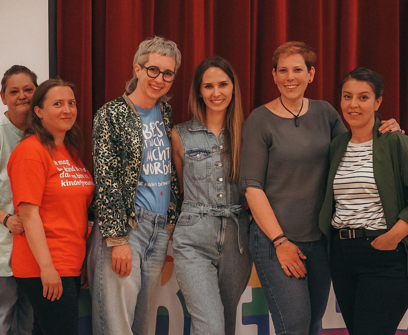 Fünf Frauen stehen lächelnd vor einem roten Vorhang. Sie tragen Freizeitkleidung. Eine trägt eine Brille, und eine andere trägt eine Halskette.