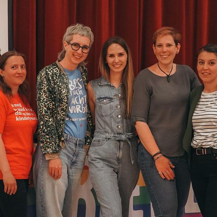 Fünf Frauen stehen lächelnd vor einem roten Vorhang. Sie tragen Freizeitkleidung. Eine trägt eine Brille, und eine andere trägt eine Halskette.
